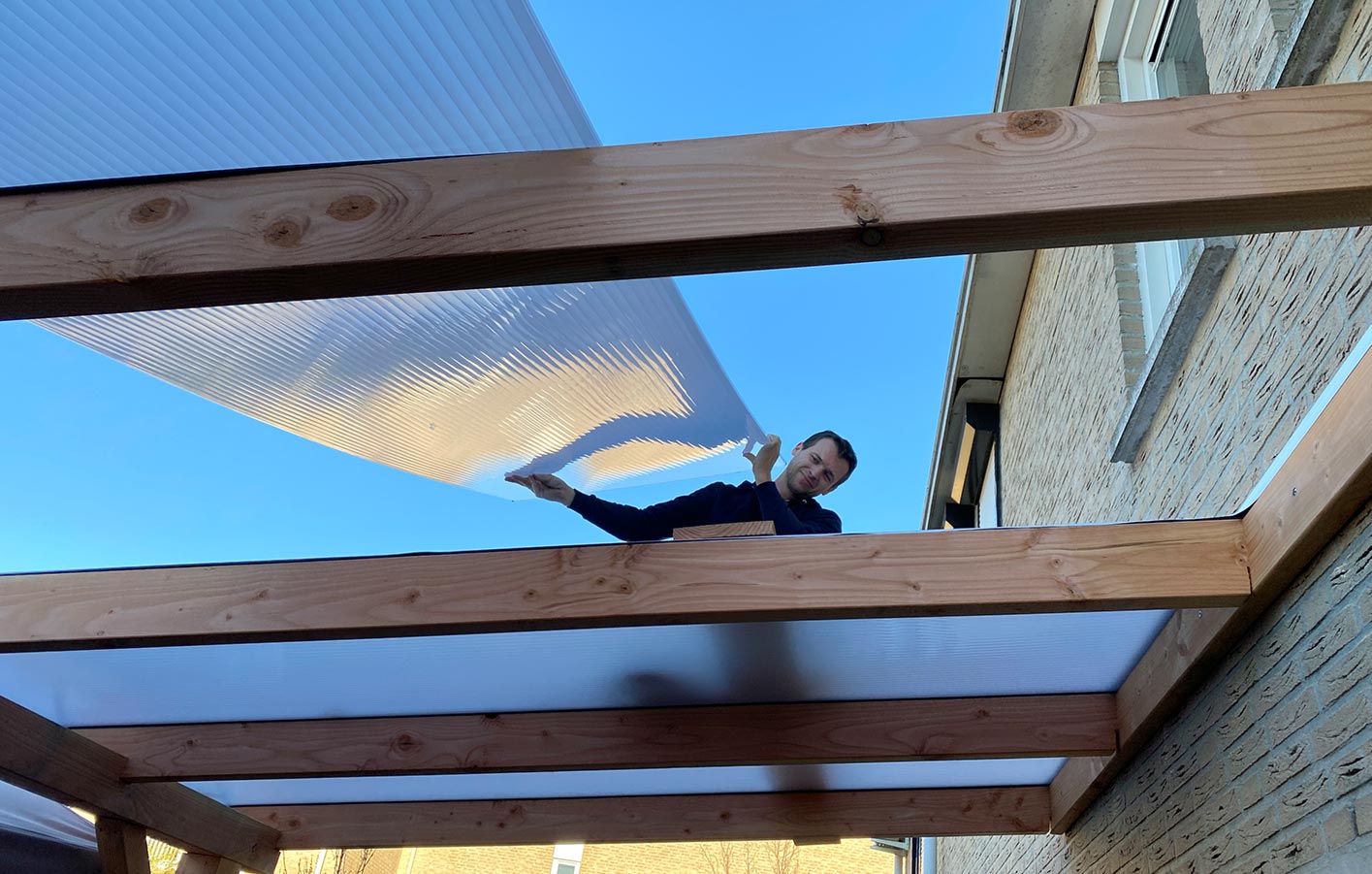 Befestigung von Polycarbonatplatten – Terrassendach Mann installiert transparente Polycarbonatplatten auf Holzbalken einer Terrassenüberdachung.