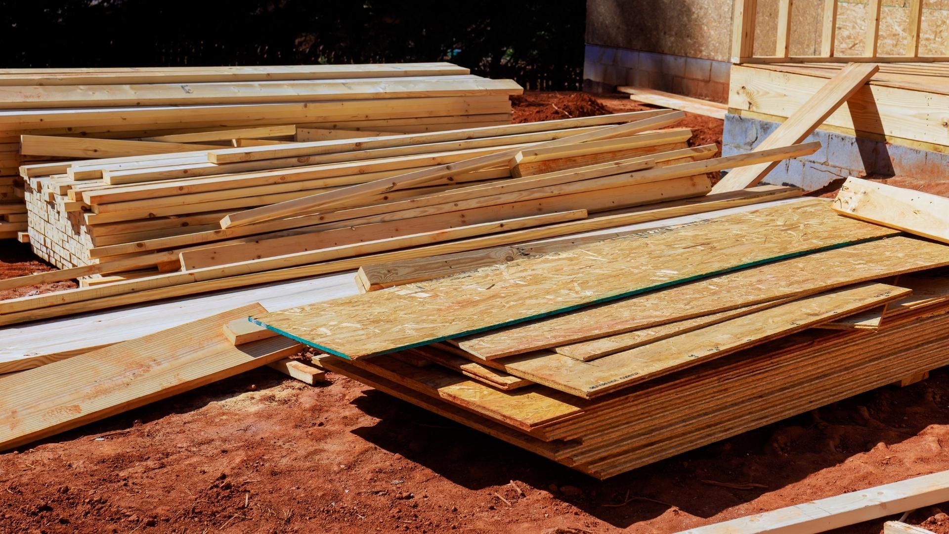 Baustellenholz ist die Grundlage für stabile und langlebige Bauprojekte. Hochwertiges Konstruktionsholz und OSB-Platten sorgen für präzise Verarbeitung, effiziente Bauabläufe und nachhaltige Ergebnisse auf jeder Baustelle. Das Bild zeigt gestapelte Holzbretter und OSB-Platten auf einer Baustelle. Im Hintergrund ist eine teilweise errichtete Holzkonstruktion zu sehen, der Boden besteht aus rötlicher Erde.