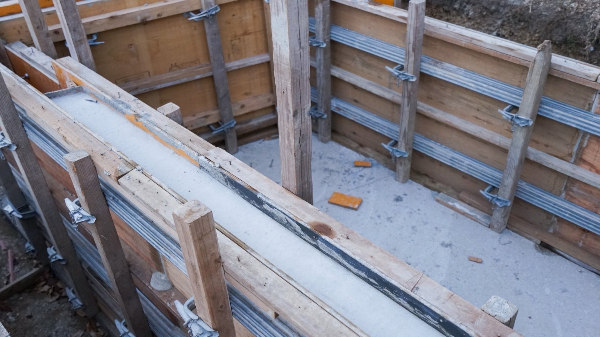 Schalungsholz ist unverzichtbar für präzise Betonarbeiten im Hoch- und Tiefbau. Mit stabilen Holzschalungen lassen sich saubere Betonflächen und exakte Formen erzielen – ideal für professionelle Bauunternehmen und Handwerker. Das Bild zeigt eine Baustelle mit montierten Holzschalungen, die mit Metallverbindern und Stützen fixiert sind. Innerhalb der Schalung ist bereits Beton gegossen, der Boden ist eben und von Bauwerkzeugen umgeben.