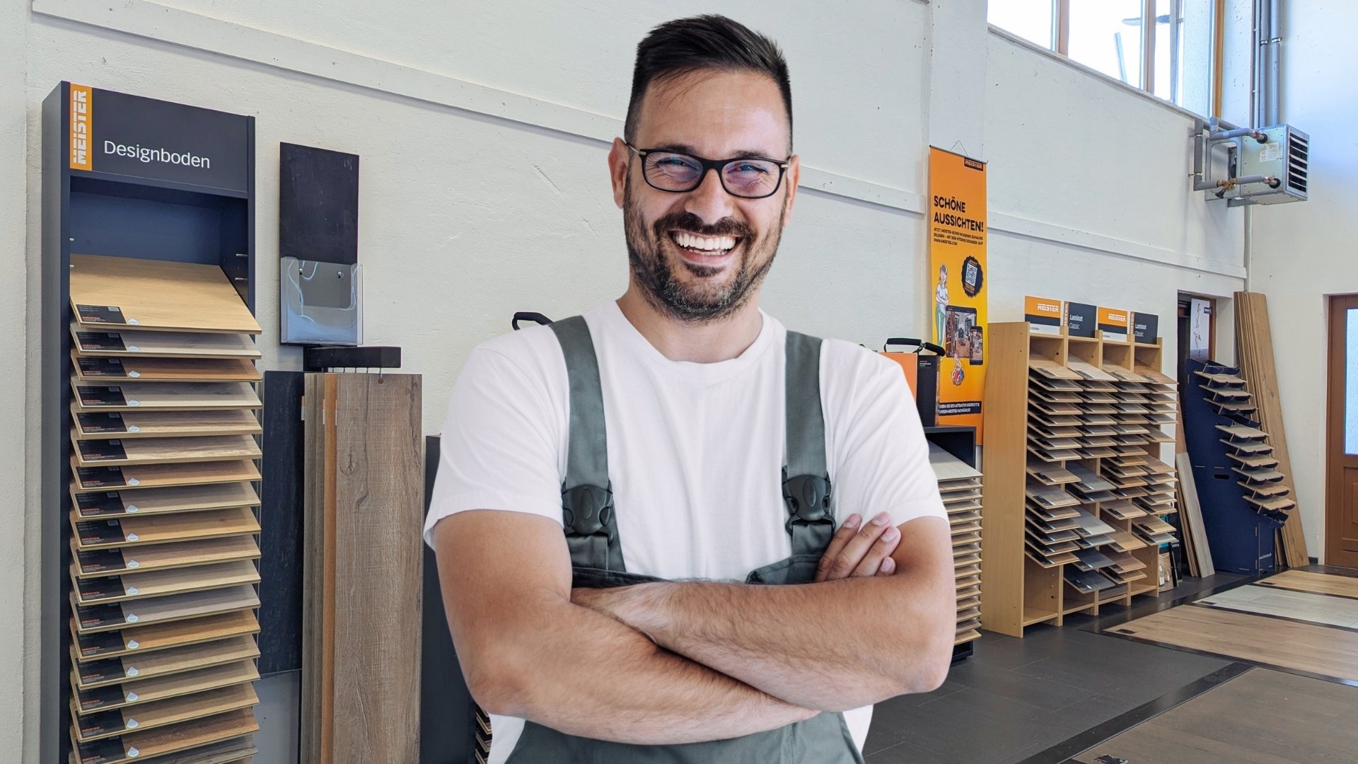 Ein Expertentipp für Laminatboden: Achten Sie auf eine hochwertige Trittschalldämmung und ein robustes Nutzungsklassen-Laminat, um sowohl Wohnkomfort als auch Langlebigkeit zu erhöhen. Eine sorgfältige Vorbereitung des Untergrunds und die Wahl passender Dekore sorgen zudem für ein rundum überzeugendes Ergebnis in jedem Raum.  Bildbeschreibung: Das Bild zeigt eine lächelnde Person in Arbeitskleidung, die mit verschränkten Armen in einer Ausstellung für Bodenbeläge steht. Im Hintergrund befinden sich Musterständer mit verschiedenen Laminat- und Designbodenmustern sowie Regale und Informationsmaterialien in einem hellen Verkaufsraum.