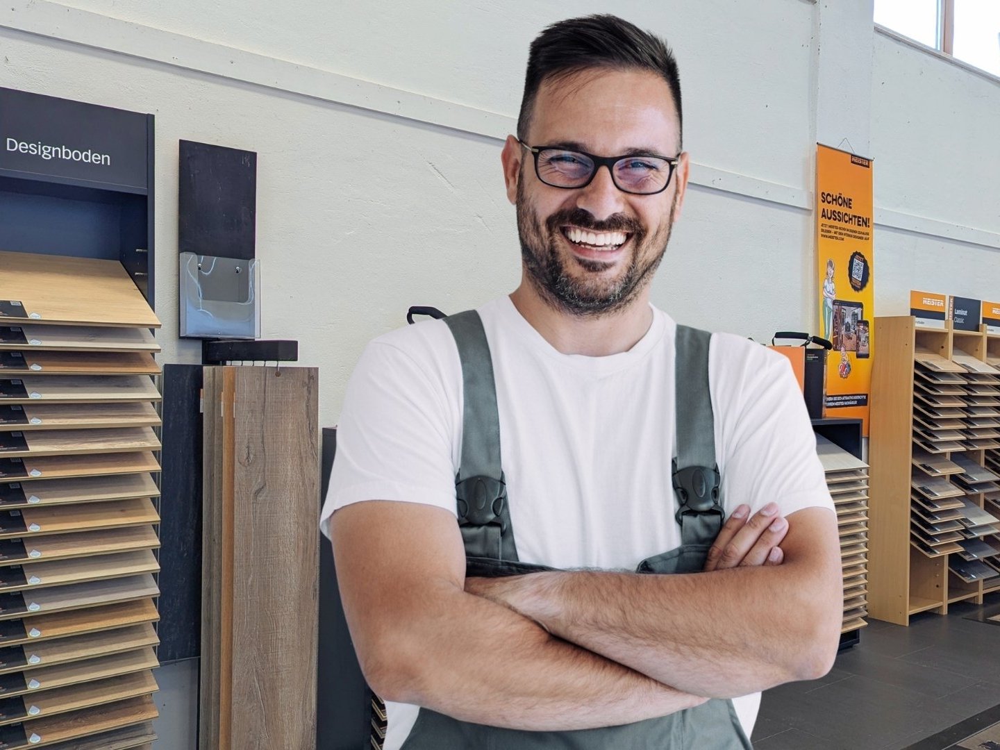 Unser Expertentipp für Linoleum und Designvinyl: Achten Sie auf eine hochwertige Materialauswahl und eine präzise fachgerechte Verlegung, um von maximaler Strapazierfähigkeit, Komfort und Designvielfalt zu profitieren. Beide Bodenarten sind ideal für moderne Wohnräume, da sie pflegeleicht, langlebig und optisch flexibel einsetzbar sind.  Bildbeschreibung: Das Bild zeigt einen lächelnden Mann in Arbeitskleidung mit verschränkten Armen, der in einer Ausstellung für Bodenbeläge steht. Hinter ihm befinden sich mehrere Musterständer mit unterschiedlichen Vinyl-, Linoleum- und Designbodenmustern sowie weitere Holzpaneele und Informationsmaterialien.