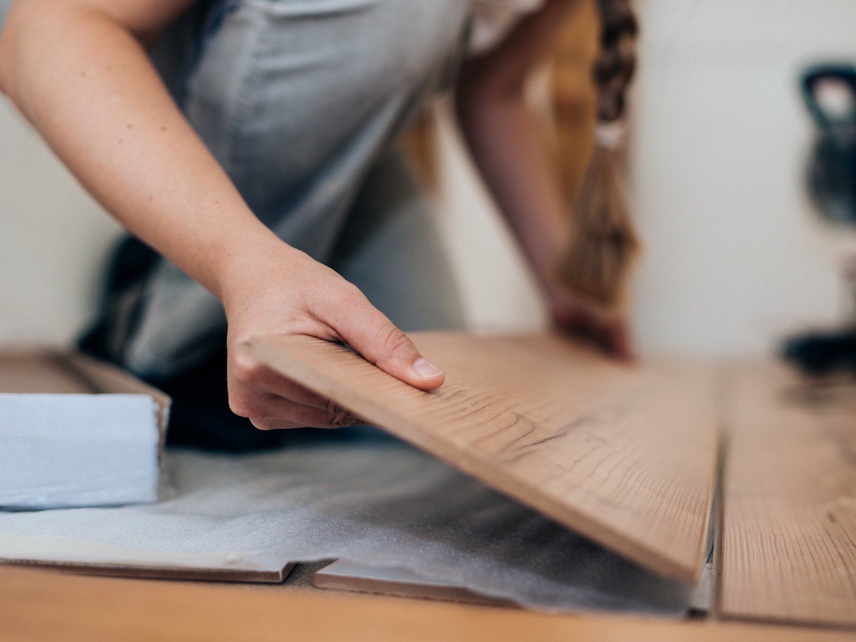 Ein Laminatboden lässt sich mit der richtigen Vorbereitung schnell und fachgerecht verlegen, sodass ein langlebiges und optisch ansprechendes Ergebnis entsteht. Dank moderner Klicksysteme gelingt die Montage auch Heimwerkern mühelos und sorgt für eine saubere, stabile und strapazierfähige Bodenfläche.  Bildbeschreibung: Das Bild zeigt eine Person, die ein Laminatpaneel in der Hand hält und es auf dem Boden positioniert, um es zu verlegen. Die Szene zeigt Nahaufnahmen der Hände, des Holzdekors der Paneele sowie eines teilweise bereits verlegten Bereichs.