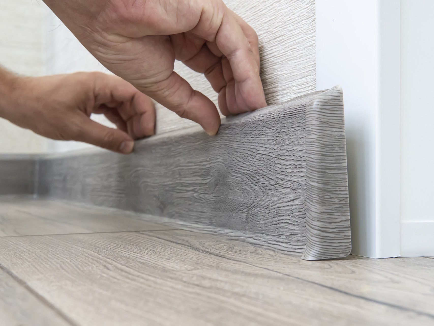 Laminat Sockelleisten und Fußleisten sorgen für einen sauberen, eleganten Übergang zwischen Boden und Wand und verleihen jedem Raum ein professionelles Finish. Mit robusten Materialien und perfekt abgestimmten Designs ergänzen sie Laminatböden optisch ideal und schützen gleichzeitig Wände und Kanten vor Abnutzung. Bildbeschreibung: Das Bild zeigt zwei Hände, die eine graue Sockelleiste in Holzoptik an einer Wand anbringen. Der darunterliegende Boden besteht aus hellem Laminat, und die Sockelleiste wird passgenau an die Wand gedrückt, um einen sauberen Abschluss zu erzeugen.