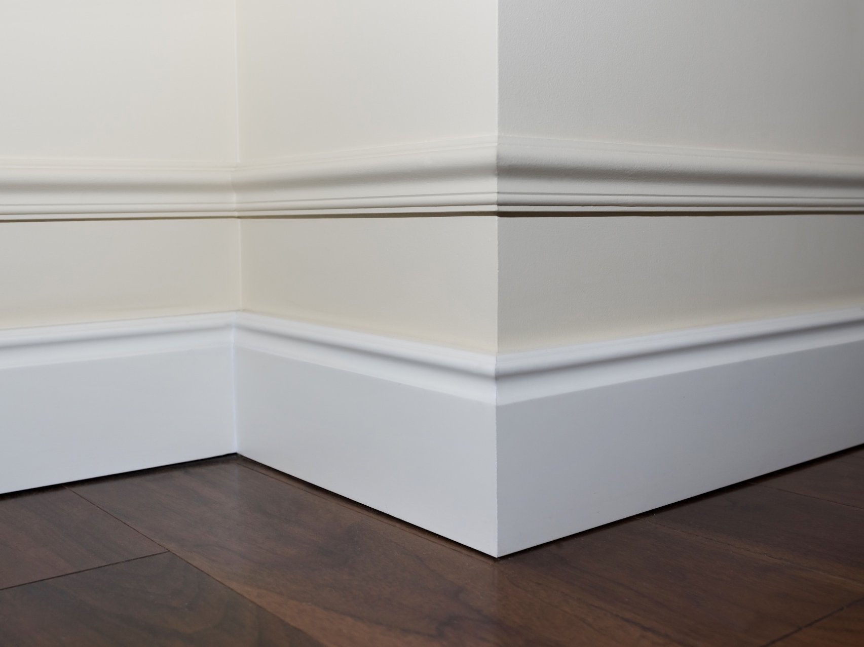 Hochwertige Fußleisten, Sockelleisten, Fußbodenkleber sowie Reiniger für Bodenbeläge runden jede professionelle Bodengestaltung optimal ab. Mit dem passenden Zubehör sorgen Sie für saubere Übergänge, eine zuverlässige Befestigung und langfristig gepflegte Bodenflächen – ideal für ein harmonisches, langlebiges Wohnambiente.  Bildbeschreibung: Das Bild zeigt eine Raumecke mit weißen Sockelleisten und dekorativen Wandprofilen. Der dunkle Holzfußboden schließt sauber an die Leisten an, wodurch ein eleganter und präziser Übergang zwischen Boden und Wand entsteht.