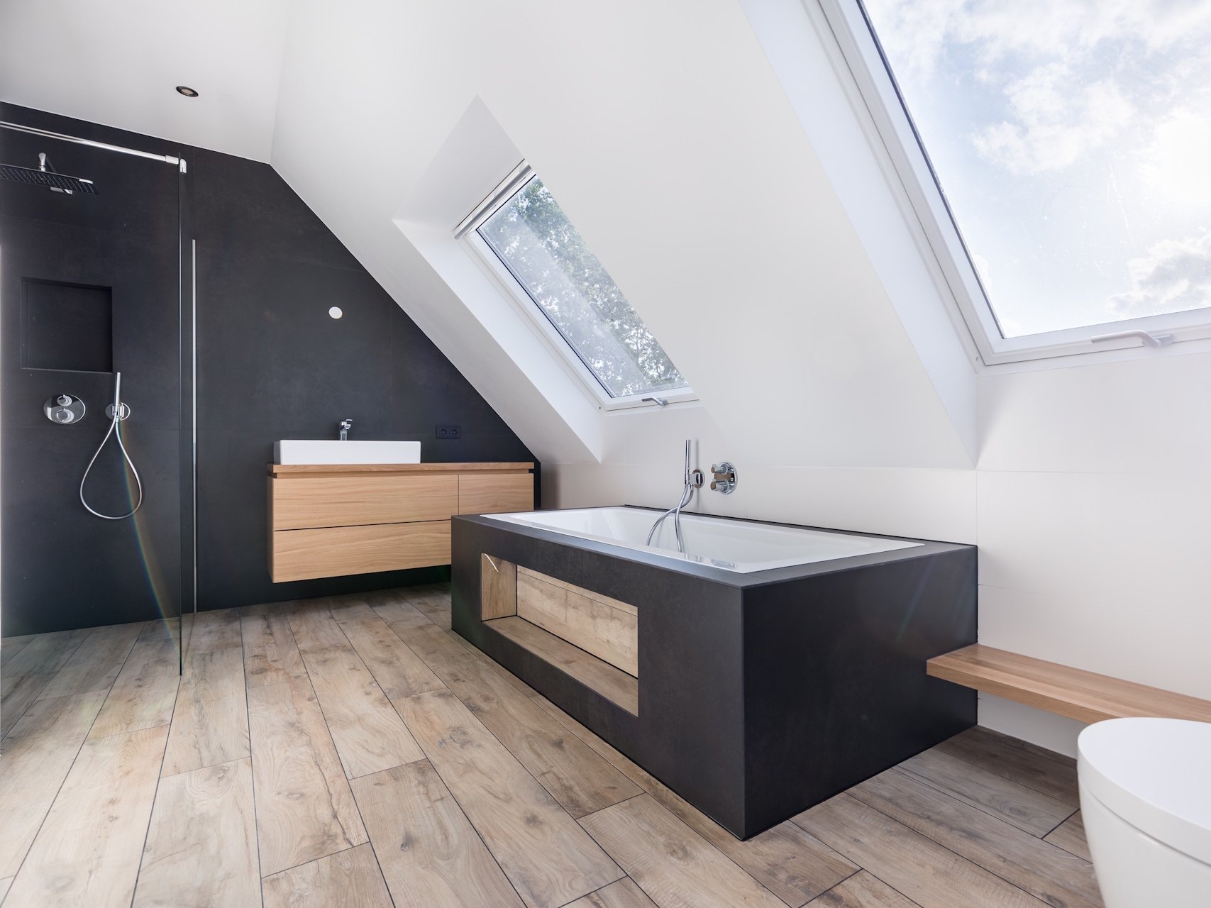 Die richtige Vinyl und Linoleum Pflege sorgt dafür, dass Ihr Boden dauerhaft schön, hygienisch und widerstandsfähig bleibt. Mit schonenden Reinigungsmitteln und regelmäßiger Pflege behalten beide Bodenarten ihre Farbintensität, Strapazierfähigkeit und ihr gepflegtes Erscheinungsbild – ideal für Feuchträume wie Bäder oder Küchen.  Bildbeschreibung: Das Bild zeigt ein modernes Badezimmer mit schrägen Decken und großen Dachfenstern, die viel Tageslicht hereinlassen. Der Raum verfügt über einen Holzoptik-Boden, eine freistehende Badewanne mit dunkler Verkleidung, eine begehbare Dusche sowie ein Waschbecken auf einem hellen Holzunterschrank.