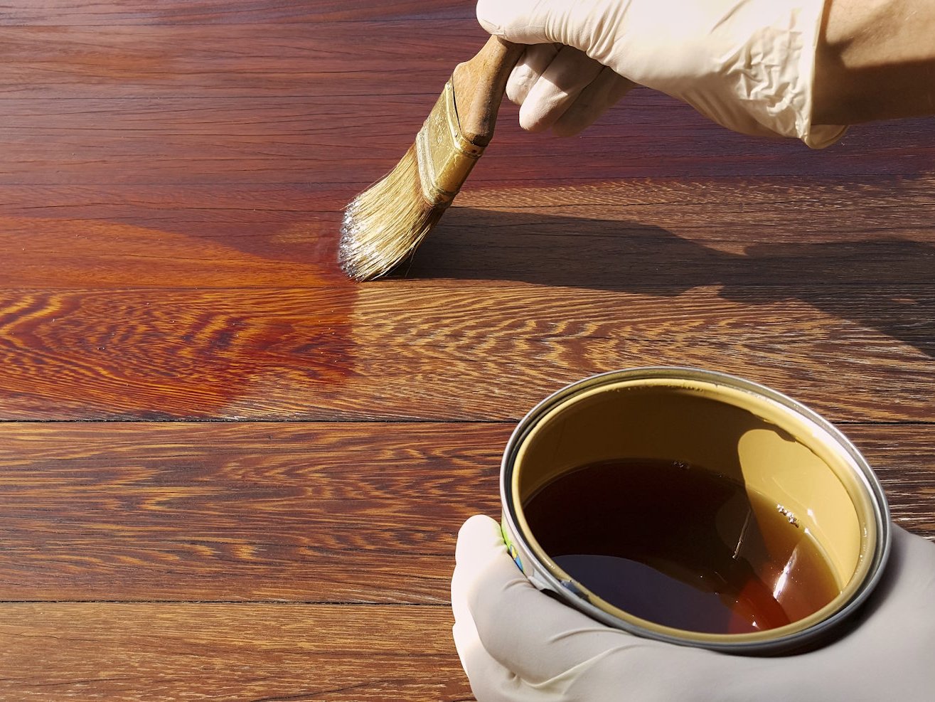 Die richtige Massivholzdielen Pflege bewahrt die natürliche Schönheit des Holzes und schützt die Oberfläche langfristig vor Abnutzung. Mit hochwertigen Pflegeölen und einer regelmäßigen Behandlung bleiben Ihre Dielen widerstandsfähig, farbintensiv und vermitteln dauerhaft ein warmes, hochwertiges Wohngefühl.  Bildbeschreibung: Das Bild zeigt zwei behandschuhte Hände, die mit einem Pinsel ein Pflegeöl auf eine dunkle Holzoberfläche auftragen. Ein deutlich sichtbarer Unterschied zwischen der geölten und der trockenen Holzfläche betont die intensive Farbvertiefung durch die Behandlung.