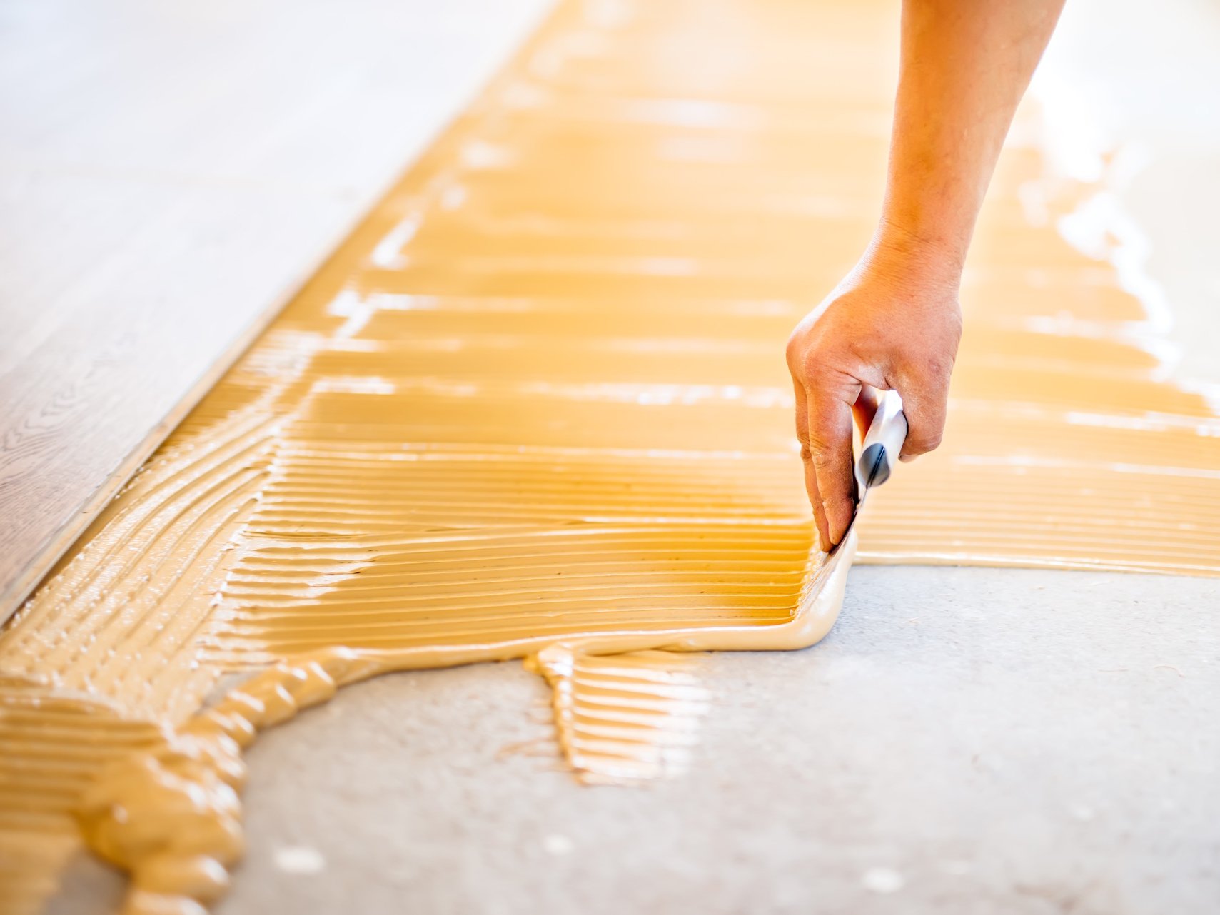 Für die professionelle Verlegung von Parkett, Vinyl, Linoleum, Kork oder Teppich ist der richtige Kleber für Bodenbeläge entscheidend. Hochwertige Bodenbelagskleber sorgen für eine dauerhaft starke Haftung, optimale Verarbeitung und ein langlebiges Ergebnis – sowohl im privaten Wohnbereich als auch in stark beanspruchten Gewerberäumen.  Bildbeschreibung: Das Bild zeigt eine Hand, die mit einer Zahnkelle einen gelblichen Bodenbelagskleber gleichmäßig auf einem Untergrund verteilt. Die strukturierten Kleberrillen und ein bereits verlegtes Paneel am oberen Rand verdeutlichen den professionellen Verlegeprozess.