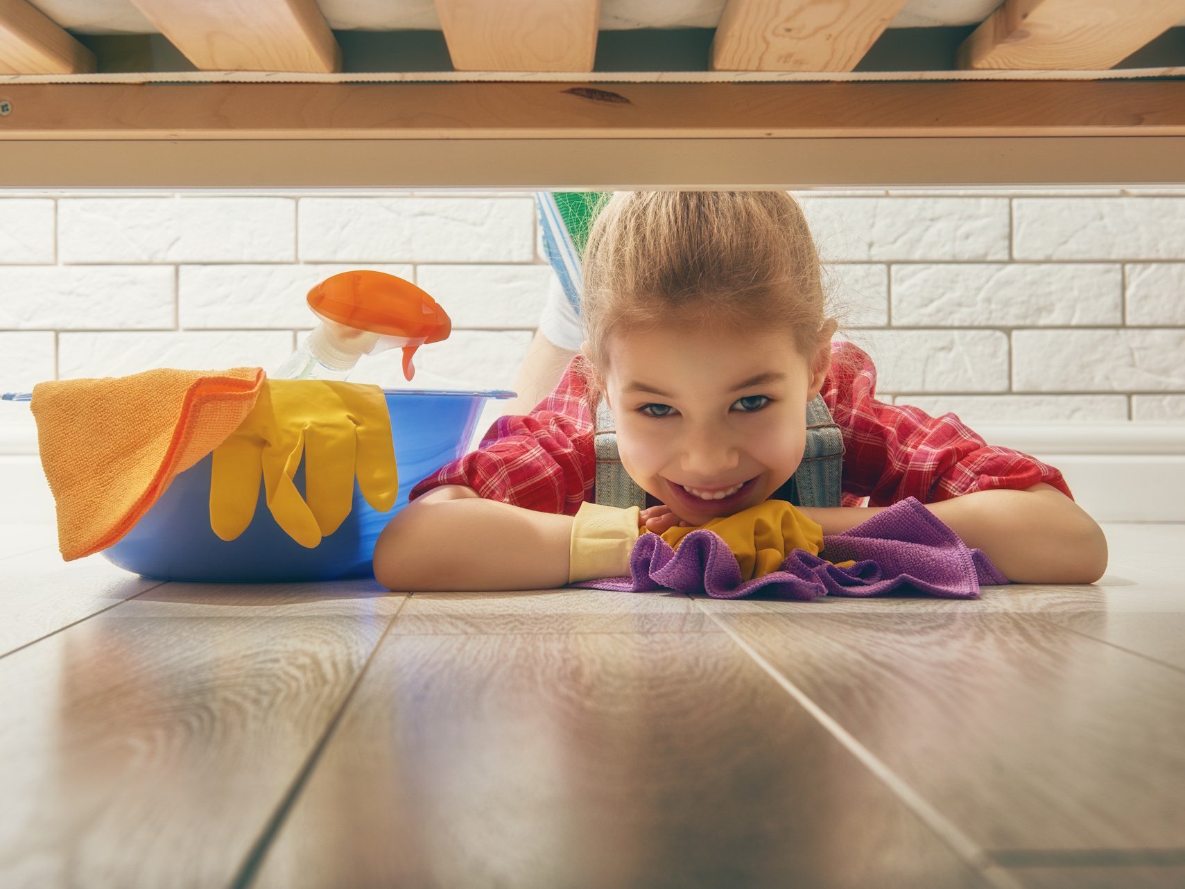Mit den passenden Pflegemitteln für Parkett, Laminat und andere Bodenbeläge bleibt Ihr Zuhause dauerhaft gepflegt und hygienisch sauber. Spezielle Reiniger schützen die Oberfläche, erhalten die natürliche Struktur Ihres Bodens und sorgen für langanhaltenden Glanz – ideal für Familien und stark genutzte Wohnbereiche.  Bildbeschreibung: Ein Kind liegt fröhlich unter einem Bett auf einem hellen Holzfußboden und wischt den Boden mit einem Tuch; daneben steht ein Eimer mit Reinigungsmitteln und Handschuhen.