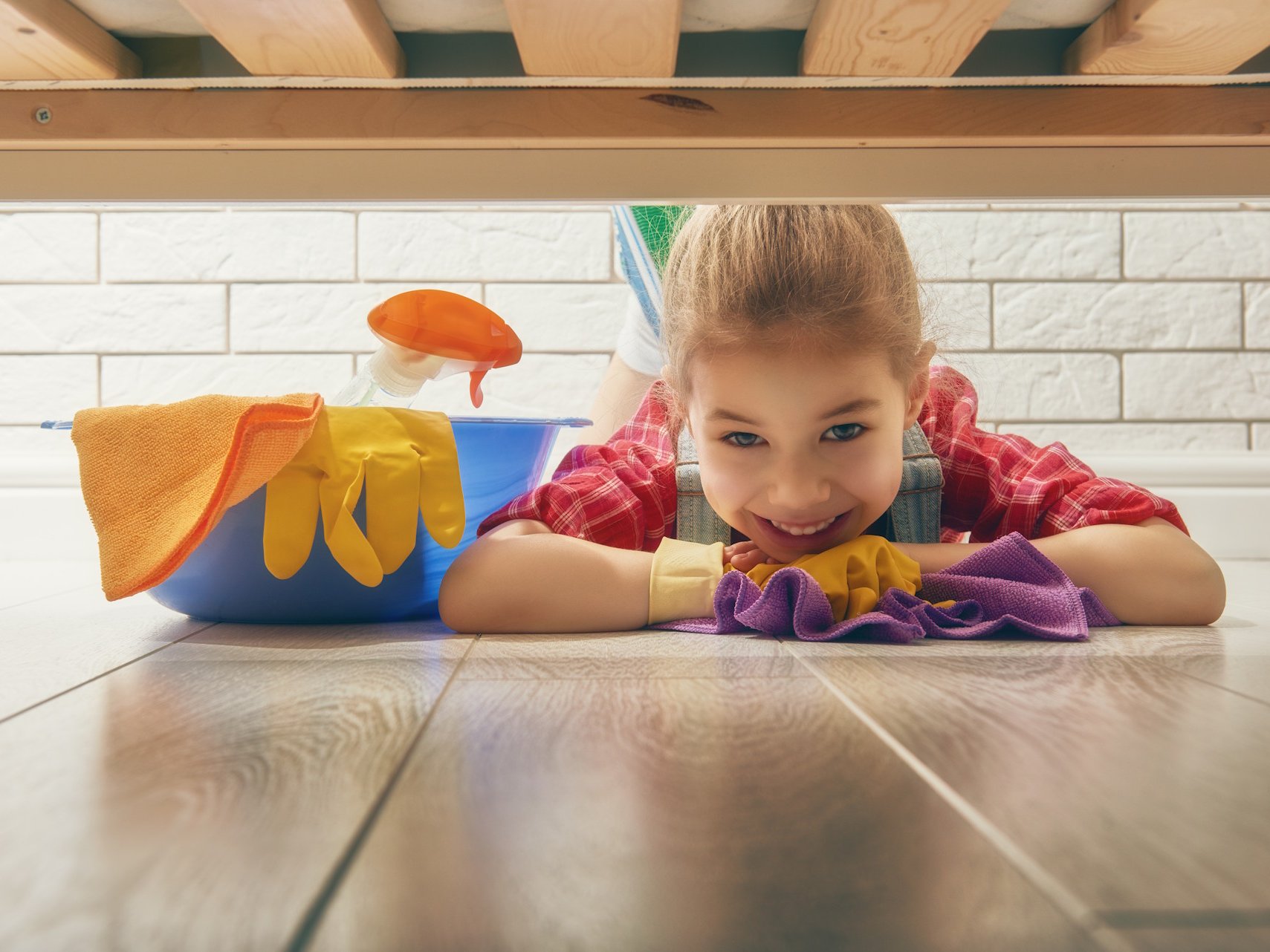 Mit den passenden Pflegemitteln für Parkett, Laminat und andere Bodenbeläge bleibt Ihr Zuhause dauerhaft gepflegt und hygienisch sauber. Spezielle Reiniger schützen die Oberfläche, erhalten die natürliche Struktur Ihres Bodens und sorgen für langanhaltenden Glanz – ideal für Familien und stark genutzte Wohnbereiche.  Bildbeschreibung: Ein Kind liegt fröhlich unter einem Bett auf einem hellen Holzfußboden und wischt den Boden mit einem Tuch; daneben steht ein Eimer mit Reinigungsmitteln und Handschuhen.