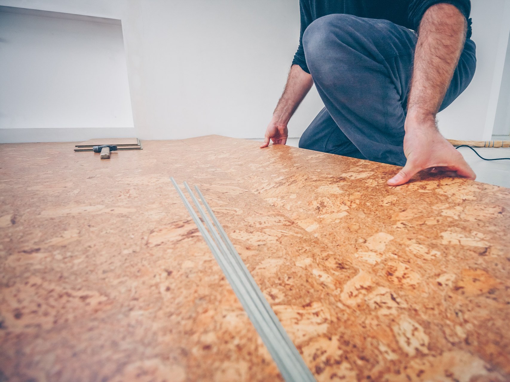 Wer Korkboden fachgerecht verlegen lässt, profitiert von einer langlebigen, elastischen und angenehm warmen Bodenfläche, die optimal in Wohn- und Arbeitsräumen funktioniert. Durch präzise Verlegung entsteht eine harmonische, fugenarme Oberfläche, die die natürlichen Vorteile von Kork voll zur Geltung bringt.  Bildbeschreibung: Das Bild zeigt eine Person, die kniend große Korkbodenplatten verlegt und sie passgenau aneinanderdrückt. Auf dem Boden liegen weitere Werkzeuge und Materialien bereit, während der noch nicht vollständig belegte Raum hell und minimalistisch wirkt.