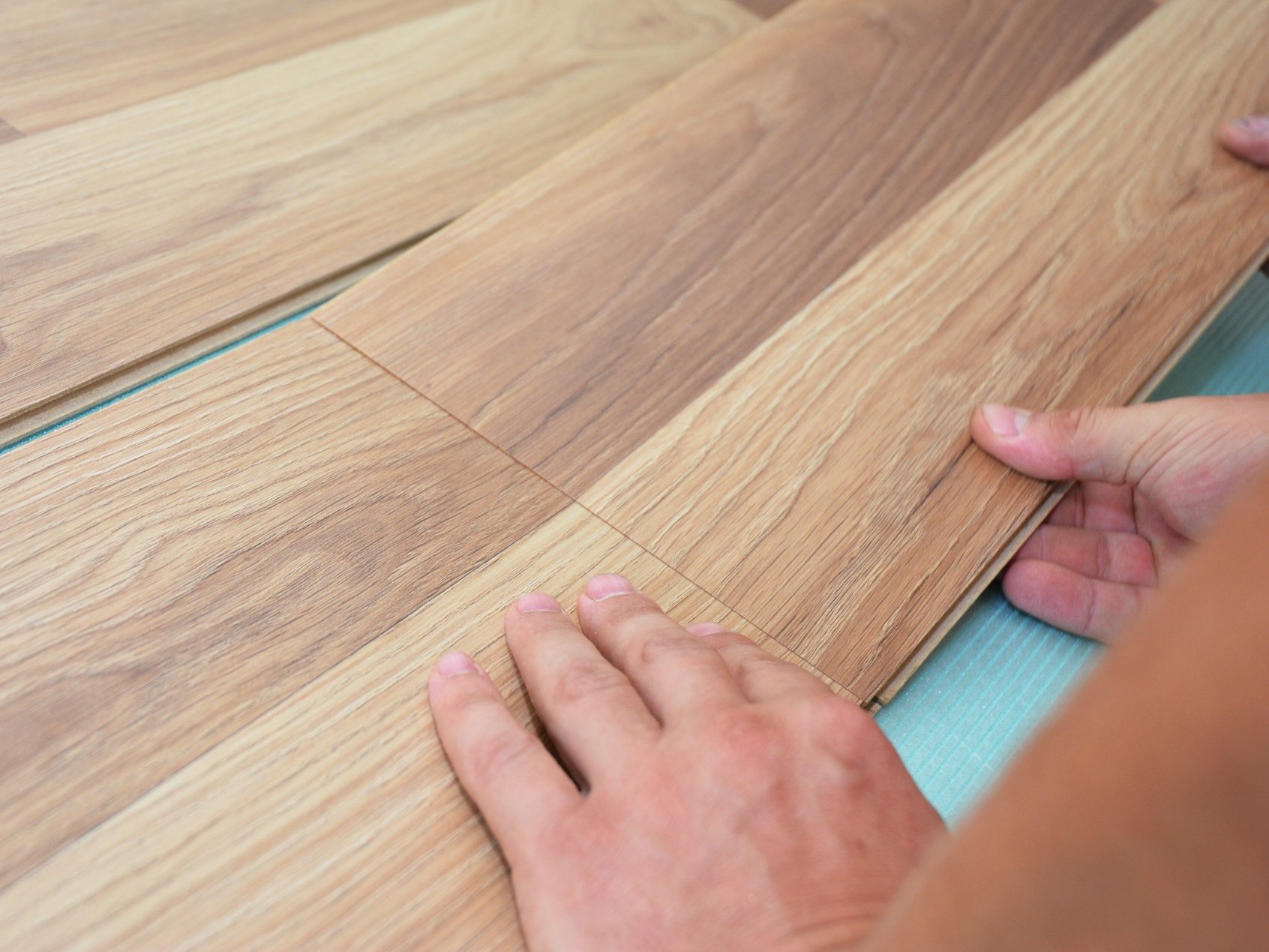 Wer Furnierboden fachgerecht verlegen möchte, profitiert von präziser Verarbeitung und hochwertigen Materialien, die für eine langlebige und optisch perfekte Bodenfläche sorgen. Durch das professionelle Verlegen entsteht eine stabile, gleichmäßige Oberfläche, die sowohl in Wohn- als auch in Arbeitsbereichen überzeugt.  Bildbeschreibung: Das Bild zeigt zwei Hände, die Holzbodenpaneele passgenau ineinander schieben, um einen Boden zu verlegen. Die Paneele haben eine natürliche Holzoptik in hellen und mittleren Brauntönen, und darunter ist eine dünne Unterlage sichtbar, auf der die Elemente montiert werden.