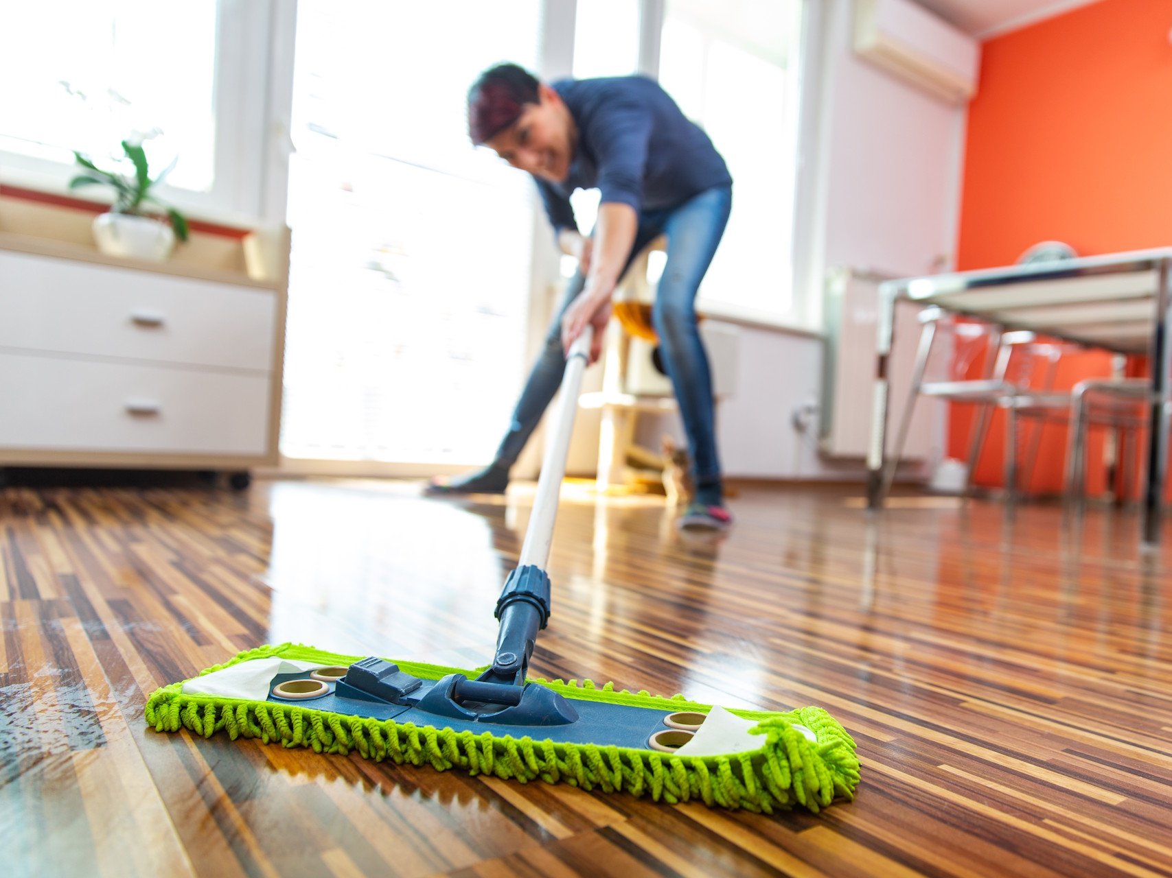 Eine regelmäßige Laminatboden Pflege sorgt für langanhaltenden Glanz und schützt die Oberfläche vor Abnutzung. Mit schonenden Reinigungsmitteln und wenig Feuchtigkeit bleibt der Boden hygienisch sauber und behält seine attraktive Holzoptik über viele Jahre hinweg.  Bildbeschreibung: Das Bild zeigt eine Person, die mit einem Wischmopp einen Laminatboden reinigt. Im Vordergrund ist der Wischmopp mit grünem Bezug zu sehen, während im Hintergrund ein heller Wohnraum mit Tisch, Stühlen und einer orangefarbenen Wand erkennbar ist.