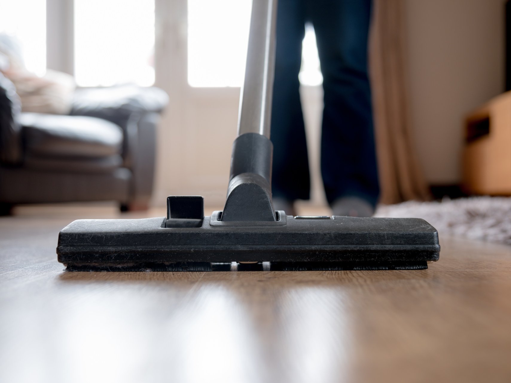 Die richtige Furnierboden Pflege sorgt dafür, dass Ihr Boden dauerhaft schön bleibt und seine natürliche Holzoptik langfristig erhält. Mit einfachen, schonenden Reinigungsmethoden und der passenden Pflege schützen Sie die Oberfläche effektiv und steigern die Lebensdauer Ihres Furnierbodens.  Bildbeschreibung: Das Bild zeigt einen Staubsauger auf einem Holzfußboden, der aus der Nahperspektive aufgenommen wurde. Im Hintergrund steht eine Person, deren Beine sichtbar sind, sowie Möbel wie ein Sofa und ein Teppich, die leicht unscharf erscheinen.