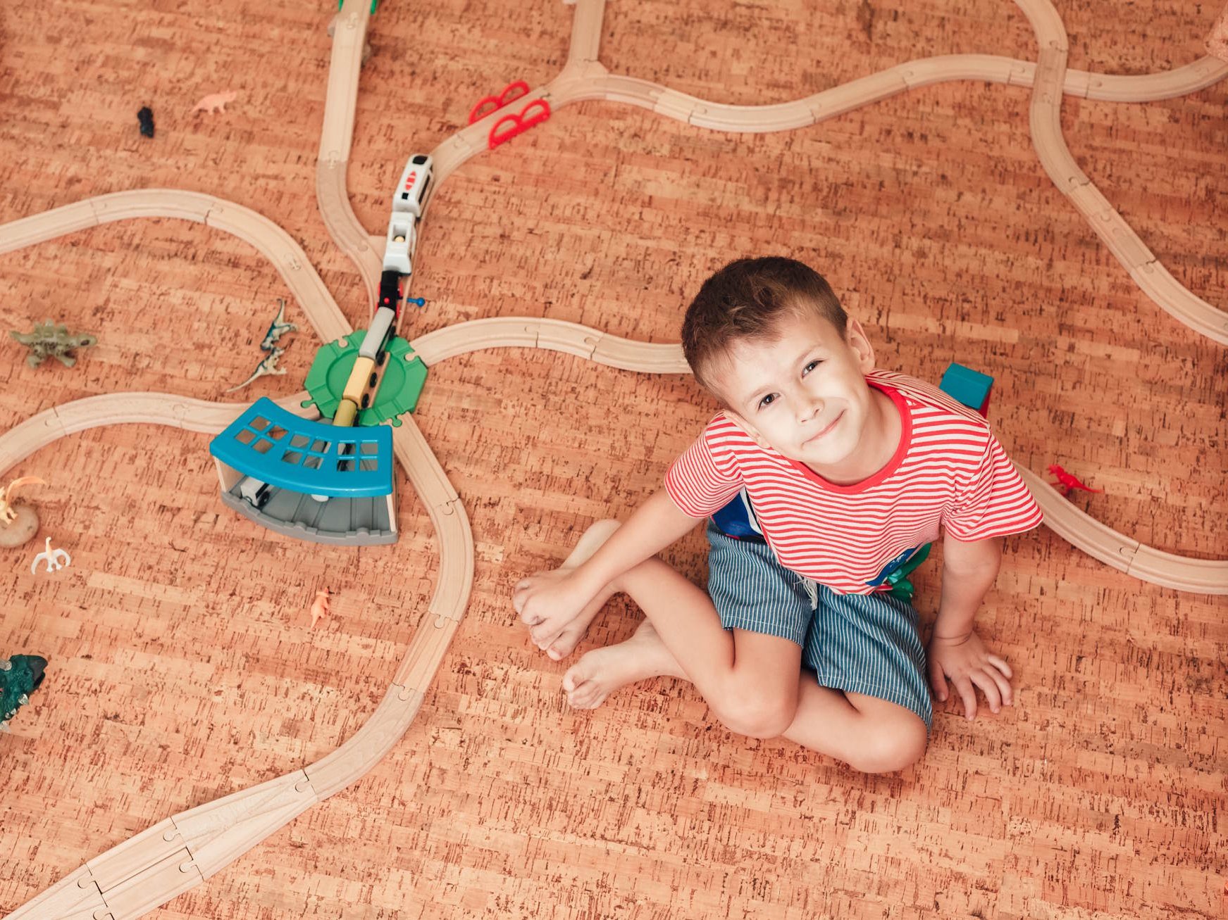 Die Korkboden Vorteile überzeugen besonders durch ihre natürliche Wärme, hohe Elastizität und die angenehm leise Raumakustik. Dank seiner fußwarmen und weichen Oberfläche eignet sich Kork ideal für Kinderzimmer und Wohnräume, in denen Komfort, Nachhaltigkeit und Wohngesundheit im Mittelpunkt stehen.  Bildbeschreibung: Das Bild zeigt ein Kind, das auf einem braunen Korkboden sitzt und inmitten einer aufgebauten Holzeisenbahn sowie verschiedener Spielzeugfiguren spielt. Die Szene wirkt warm, freundlich und zeigt den Boden als weiche, angenehme Spielfläche.